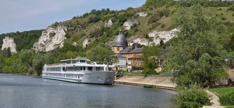       River cruise ship moored beside cliff-side village with domed chapel on the Seine.
  