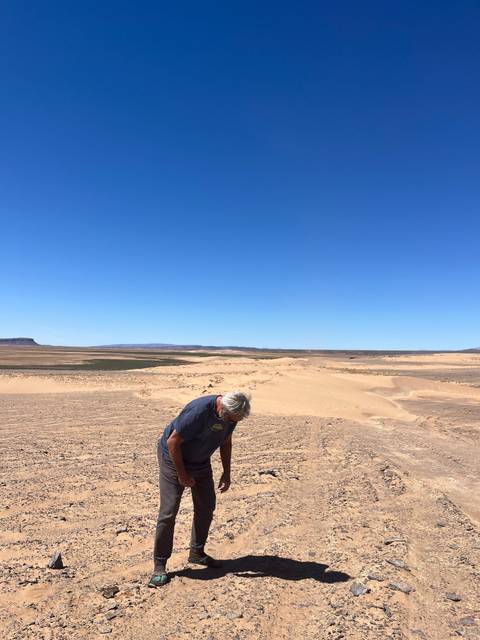       Older man examining desert soil under a bright blue sky and flat horizon.
  