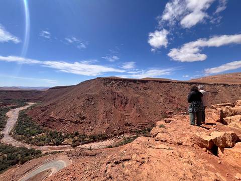       Two people stand on red rock cliff above winding river valley in the desert.
  