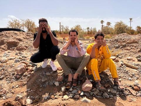       Three friends mimic see-no-evil pose while sitting on rocky desert ground.
  