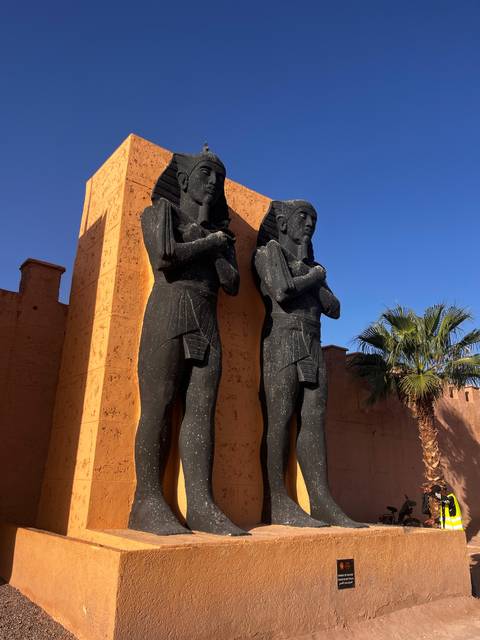       Two large black Egyptian-style statues stand against an ochre wall with a palm tree and clear blue sky.
  
