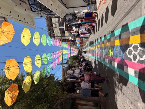       Bustling market street under a canopy of bright umbrellas and multicolored painted walkway.
  