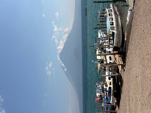       Clear view of small boats moored at a lakeside pier with a prominent volcano rising across turquoise waters under a blue sky.
  