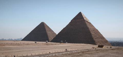       Two monumental pyramids rise from the sandy Giza plateau with Cairo’s skyline faint on the horizon.
  