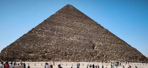       The Great Pyramid of Giza towers over clusters of tourists exploring its massive stone base.
  