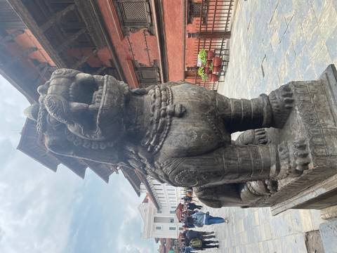       Stone guardian lion statue in Kathmandu Durbar Square with tourists exploring historic brick palaces.
  