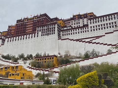       Potala Palace’s massive white and ochre walls cascade down the hillside under a cloudy Tibetan sky.
  