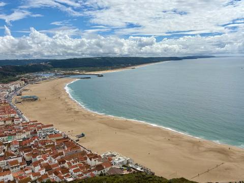       Expansive aerial view of Nazaré’s long sandy beach meeting the calm Atlantic under a partly cloudy sky.
  
