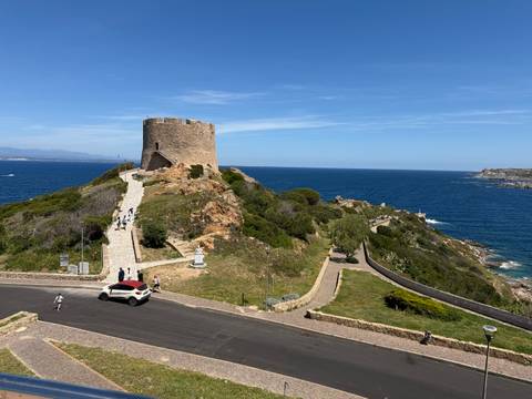       Stone coastal watchtower on a rocky promontory overlooking deep blue Mediterranean waters with visitors walking the path.
  