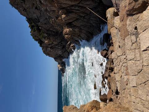       Waves crash against jagged granite rocks on a rugged Sardinian shoreline under a clear sky.
  