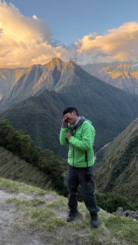       Young trekker in green jacket laughs with Machu Picchu terraces and steep mountains behind.
  