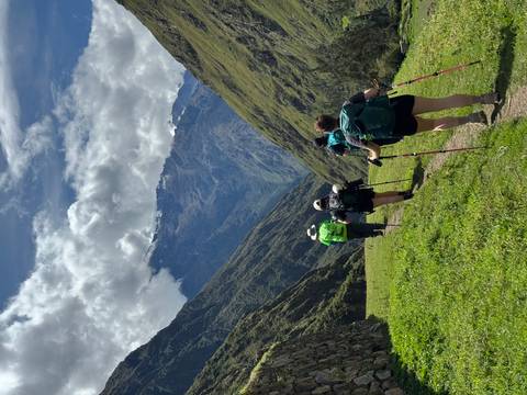       Line of hikers with trekking poles strides along a green mountain valley trail toward snow-capped summits.
  