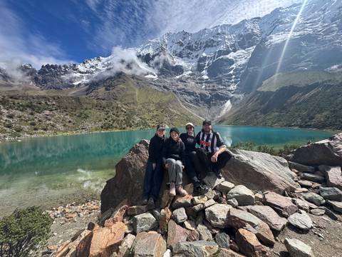       Four travelers sitting on rocks by a vibrant emerald lake with snowy peaks above.
  
