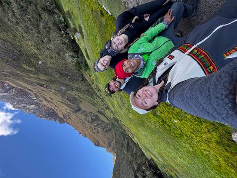       Friends taking a wide-angle selfie in a mossy alpine meadow beneath steep cliffs.
  