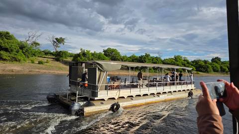       Flat-bottomed safari boat cruising a river while passengers observe the shoreline; hands with phone capture the scene in foreground.
  