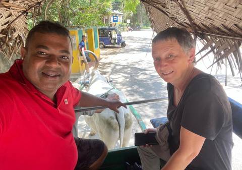       Guide and tourist smile for selfie while riding in a shaded bullock cart along leafy road in Sri Lanka.
  