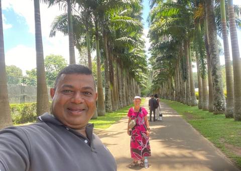       Traveller stands on palm-lined avenue while guide takes selfie foreground; manicured gardens around.
  