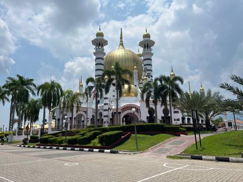       Majestic Ubudiah Mosque with golden dome and palm trees against a partly cloudy sky.
  