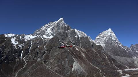       Red helicopter soars past jagged, snow-dusted Himalayan peaks under a deep blue sky.
  