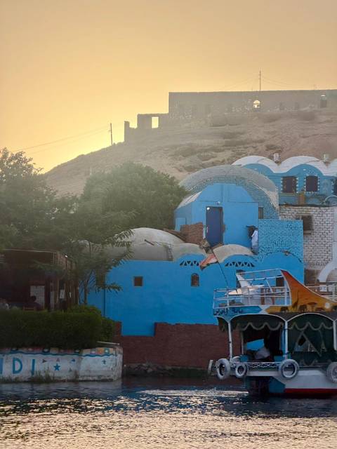       Sun-washed view of blue-painted domed Nubian houses nestled against a rocky hillside beside the Nile.
  