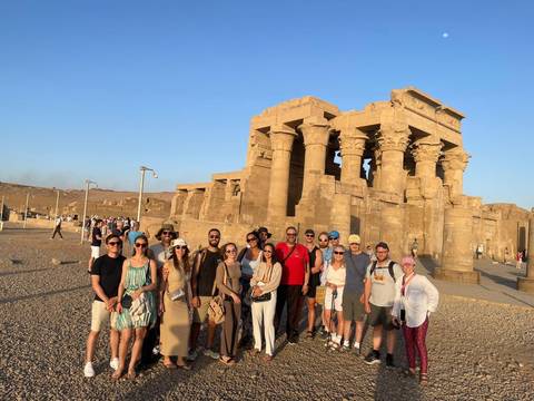       Large tour group standing before the massive pillars of Kom Ombo Temple in warm evening light.
  