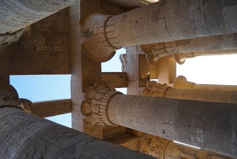       Upward view of ornately carved capitals and cross-beams of an ancient Egyptian temple against a bright sky.
  