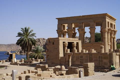       The small kiosk structure of Philae Temple stands beside the Nile with palm trees and rocky outcrops.
  