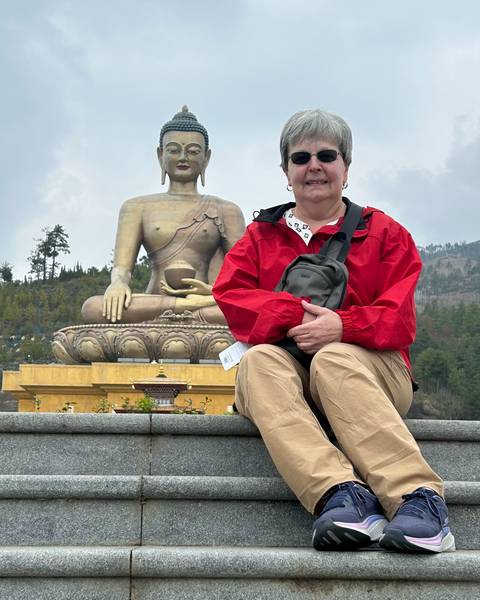       Smiling traveler sitting in front of the giant golden Buddha Dordenma statue on a cloudy day.
  
