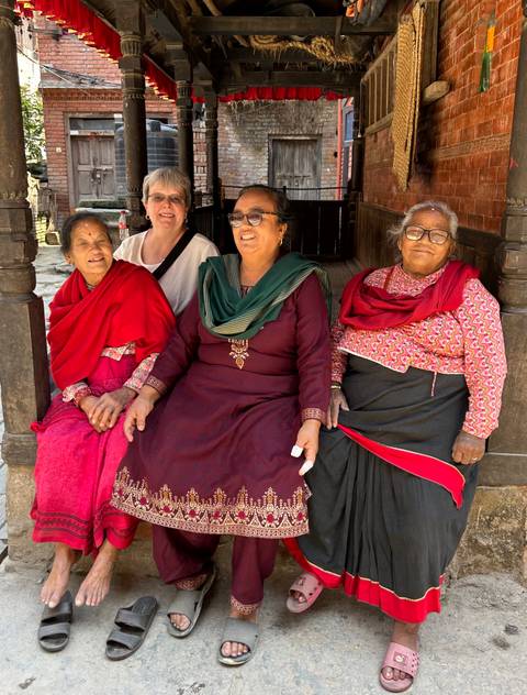       Tourist sitting with three elderly women in bright traditional clothing outside a wooden building.
  