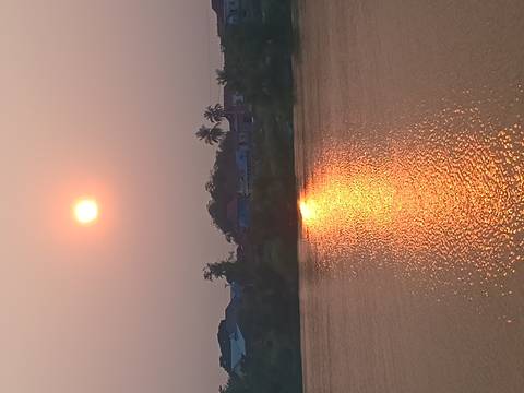       Orange sun sets above a riverside village, its reflection shimmering on calm water.
  