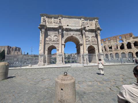       Arch of Constantine with partial Colosseum in background on a bright clear day, tourist nearby
  
