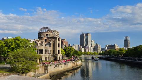       The Hiroshima Atomic Bomb Dome stands beside a river with a modern city skyline under a clear blue sky.
  