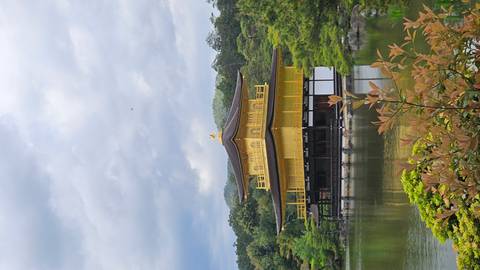       The golden Kinkaku-ji pavilion shimmers above a reflective pond surrounded by lush greenery and moody clouds.
  