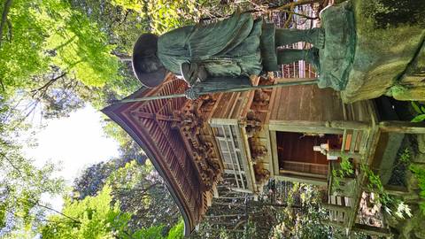       A small wooden shrine and a bronze monk statue nestle in a lush forest, lit by dappled sunlight.
  