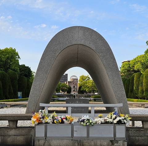       The arched cenotaph frames the Atomic Bomb Dome in Hiroshima Peace Memorial Park amid manicured greenery.
  