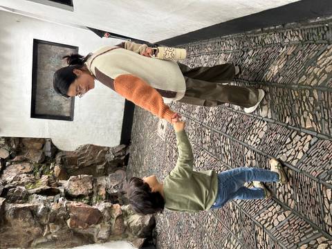       Woman holding a young child’s hand while walking along a cobblestone pathway near rock walls
  