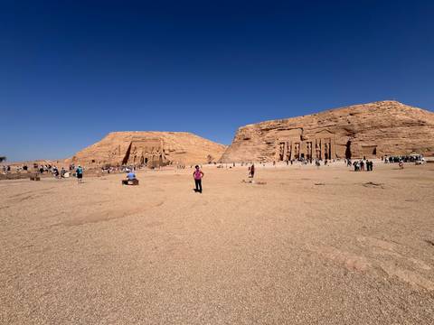       Wide view of the Abu Simbel temples carved into a desert cliff with visitors scattered across the forecourt.
  