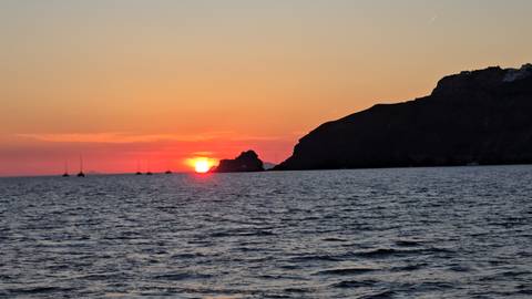       Orange sun setting behind a dark island silhouette with small sailboats on the sea.
  