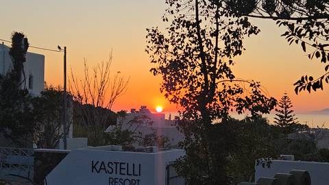       Sunset seen through silhouetted trees and resort buildings with the sign 'Kastelli' in view.
  