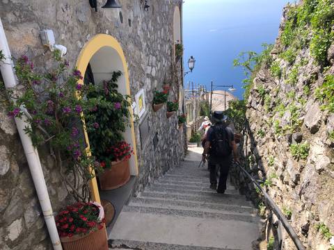       Travelers descend a narrow stone staircase lined with flowers between cliffside houses overlooking the Amalfi Coast
  