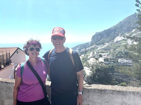       Smiling couple with backpacks pose on a terrace overlooking terraced hills and the Amalfi coastline
  
