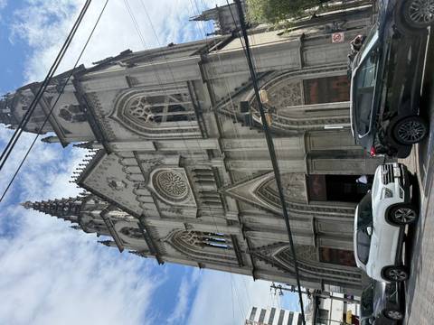       Gothic-style stone basilica with pointed spires seen against a bright blue sky, cars parked below.
  