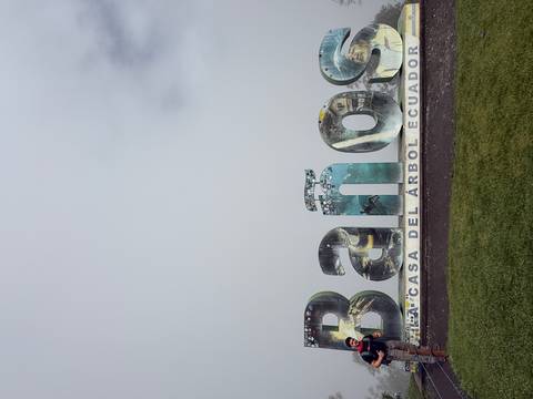       Large sculpted letters spelling Baños with travel photos embedded, set against thick mountain mist.
  
