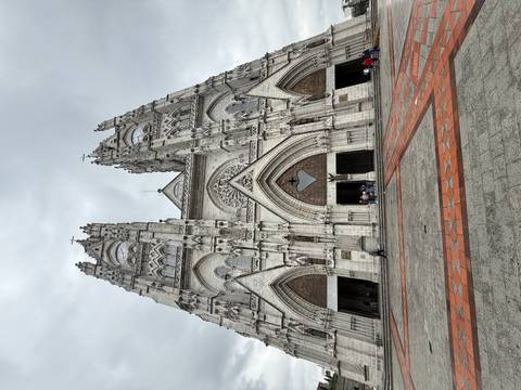       Full façade view of a soaring grey stone basilica with visitors entering beneath pointed arches.
  