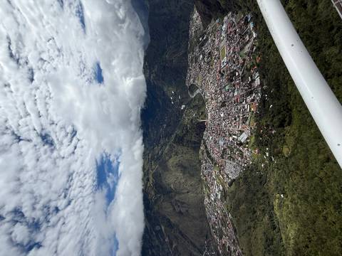       High aerial view of Baños town sprawling in a green valley beneath cloud-capped mountains.
  