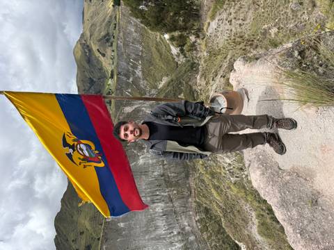       Traveler stands by an Ecuador flag atop a dramatic crater rim with steep green walls behind.
  
