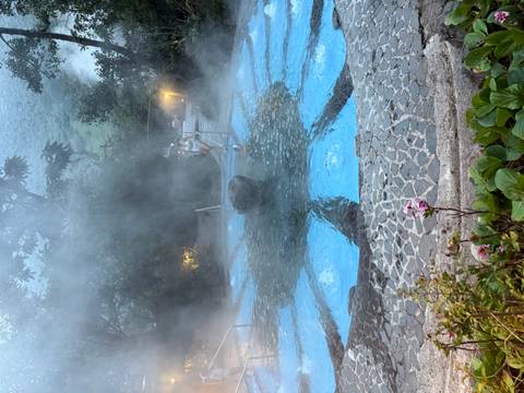       Steaming blue-tiled thermal pool at dusk with bathers enjoying the warm water in a lush setting.
  