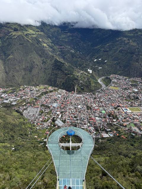       Bird’s-eye view of Baños town with surrounding green mountains and a circular sky swing platform in foreground.
  