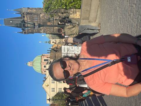       Female tourist wearing headset standing on the Charles Bridge with Prague’s domes and towers behind.
  