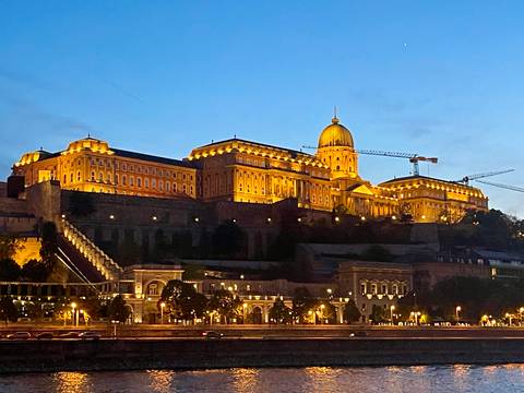       Night view of Buda Castle complex illuminated in golden light above the Danube River.
  